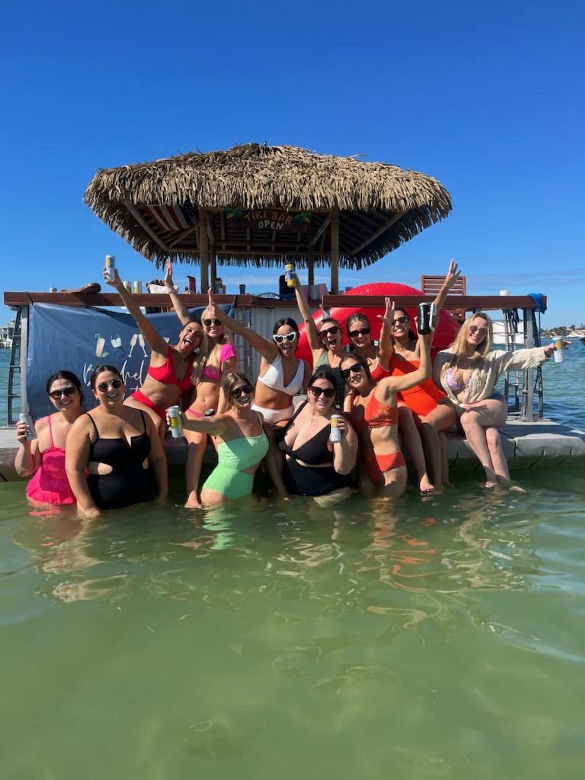 Group of friends in colorful swimwear cheering on a floating tiki-bar dock in shallow green water with a clear blue sky and boats in the background, sunny tropical beach/lagoon vibe.