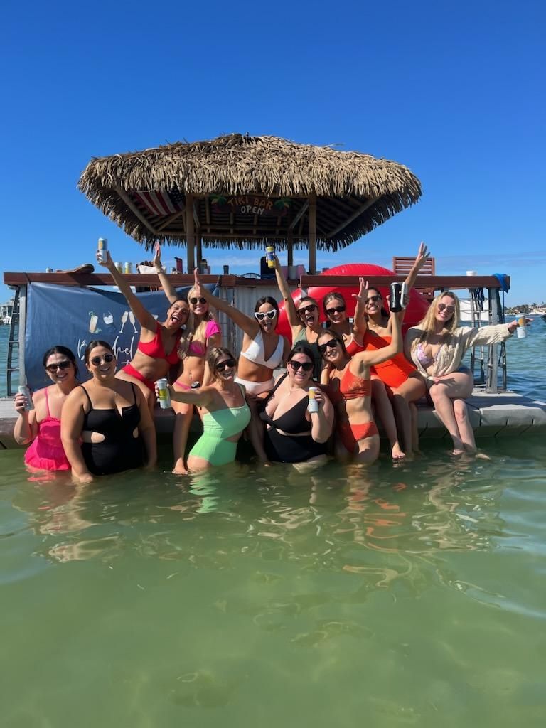 Group of friends in colorful swimsuits cheering with drinks while standing and sitting in shallow green water by a thatched-roof floating tiki bar and dock on a sunny blue-sky day.