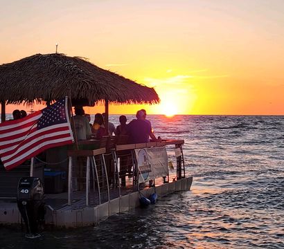 Silhouetted group on a thatched-roof floating tiki bar boat with an American flag, watching a golden ocean sunset over calm coastal waters
