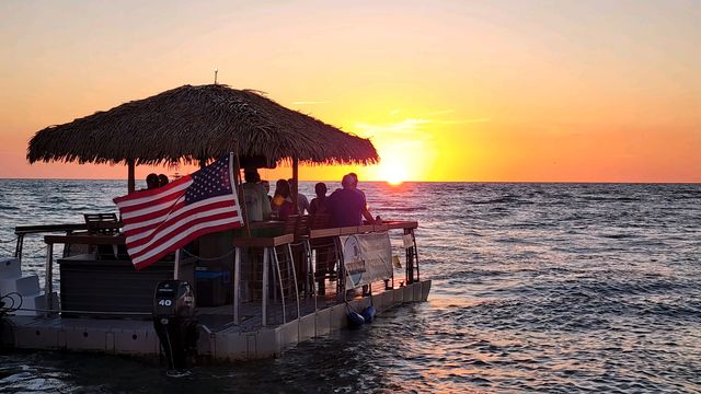 Silhouetted group on a thatched-roof floating tiki bar boat with an American flag, watching a golden ocean sunset over calm coastal waters
