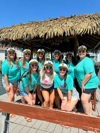 Smiling women in matching teal tees and captain hats wearing heart-shaped sunglasses pose under a thatched tiki bar on a sunny waterfront dock for a bachelorette celebration.