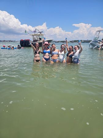 Six friends in swimsuits raising drinks on a sunlit sandbar, standing waist‑deep in shallow coastal water with anchored boats and a bright blue summer sky.