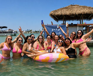 Laughing group of women in colorful swimsuits celebrating a bachelorette party on a pastel inflatable in shallow turquoise coastal bay by a tiki hut and boats under a clear blue sky