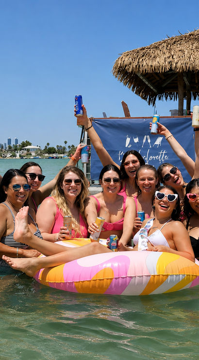 Laughing group of women in colorful swimsuits celebrating a bachelorette party on a pastel inflatable in shallow turquoise coastal bay by a tiki hut and boats under a clear blue sky