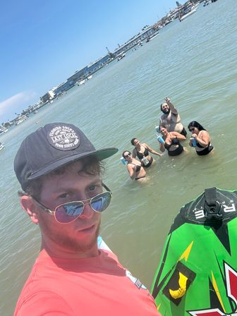 Selfie of a person in sunglasses and a cap on a green inflatable near a sunny coastal bay, five friends waist‑deep in the water holding drinks and waving, with boats and waterfront buildings in the background.