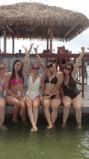 Five women in swimsuits sitting on a floating tiki bar platform with a 'Bachelorette Party' banner, feet dangling in shallow coastal water under a thatched roof.