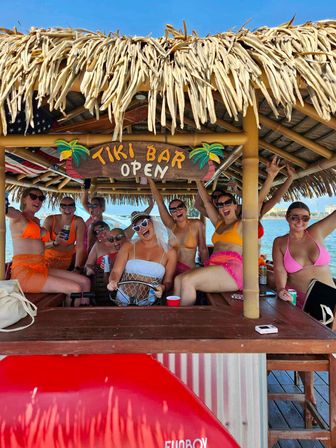 Group of friends in colorful swimsuits celebrating on a floating tiki bar with a thatched roof and Tiki Bar Open sign over blue water — summer boat party