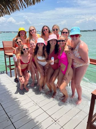 Group of women in colorful swimsuits and bucket hats posing on a sunny tropical dock over turquoise bay water