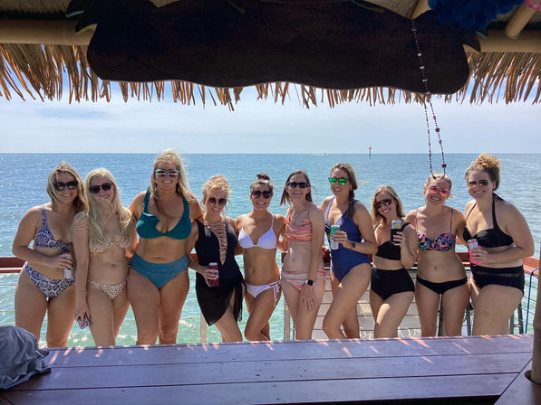 Group of friends in colorful swimsuits posing with drinks under a thatched-roof beach bar, turquoise ocean and clear sky in the background.