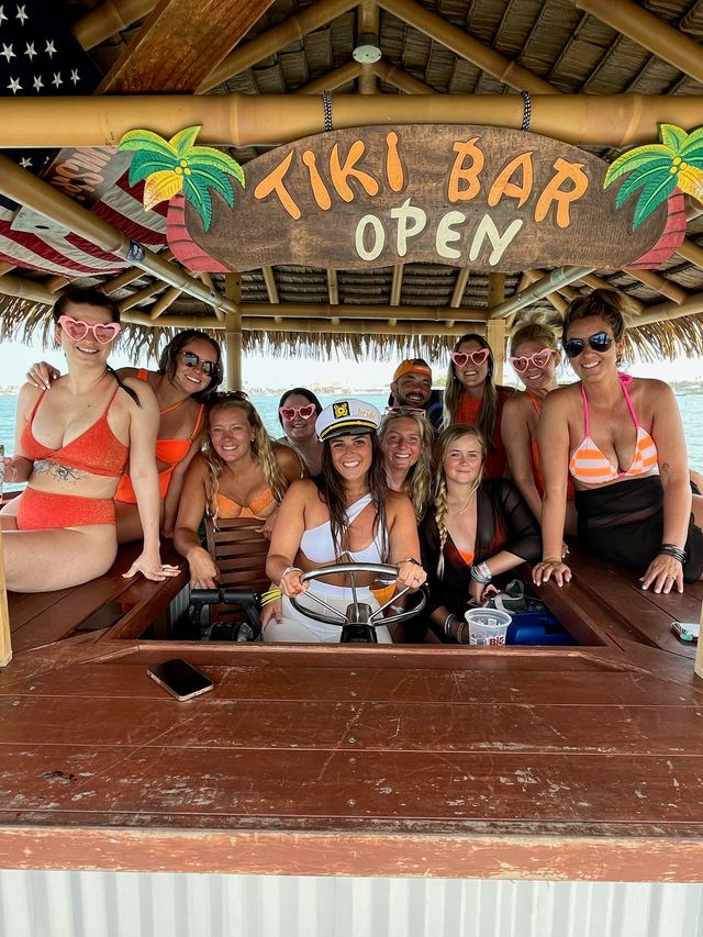 Smiling group of friends in colorful swimsuits and heart-shaped sunglasses posing under a 'TIKI BAR OPEN' thatched-roof sign on a tiki boat over a sunny coastal bay, woman at the helm wearing a captain hat