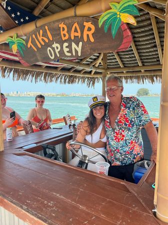 Sunny waterfront tiki bar on a boat — smiling people in swimwear at the helm under a “Tiki Bar Open” sign, woman wearing a captain’s “bride” hat and man in a floral shirt with blue bay and shoreline behind them.