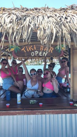 Group of friends in colorful bikinis posing and laughing under a thatched-roof “Tiki Bar Open” sign aboard a waterfront tiki bar boat on a sunny day