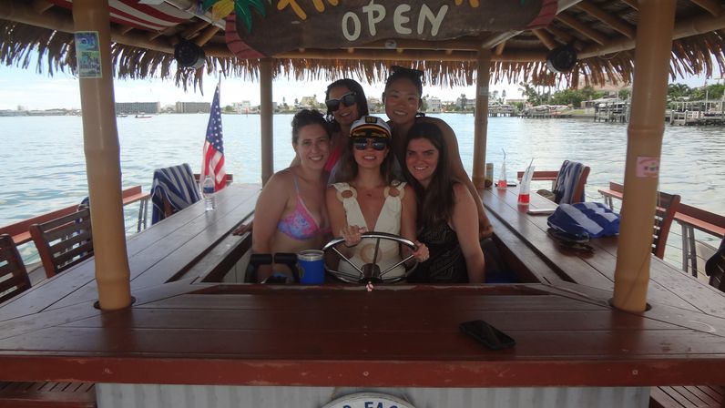 Five women smiling at the helm of a tiki-style floating barboat, hands on the captain’s wheel with an American flag, waterfront docks and calm bay in the background on a sunny day.