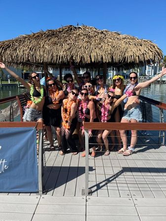 Women in swimsuits and colorful leis posing cheerfully under a thatched tiki hut on a sunny marina dock — playful girls' trip celebration.