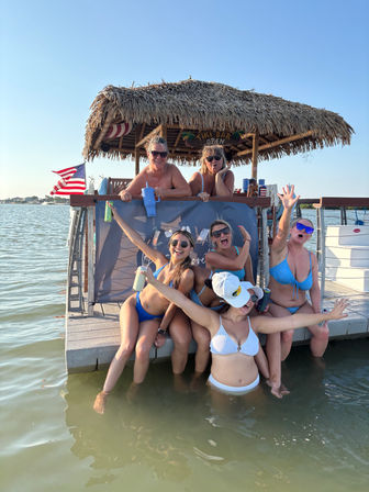 Group of friends in bikinis cheering and posing at a thatched-roof tiki bar on a sunny dock with feet in shallow bay water, holding drinks and an American flag in the background