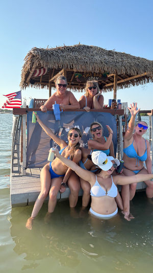 Group of friends in bikinis cheering and posing at a thatched-roof tiki bar on a sunny dock with feet in shallow bay water, holding drinks and an American flag in the background