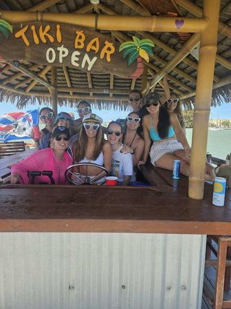 Smiling group of friends in summer outfits and heart-shaped sunglasses gathered under a thatched waterside tiki bar sign reading "TIKI BAR OPEN," with a steering wheel, drinks, palm-roof and ocean in the background.