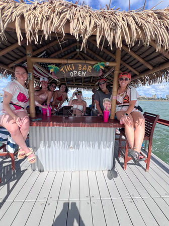 Group of friends in swimsuits smiling under a thatched-roof tiki bar on a sunny waterfront dock, holding colorful cups and sunglasses with a 'Tiki Bar Open' sign.
