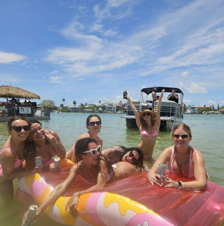Group of friends in bikinis lounging on a colorful inflatable float in a sunny shallow coastal bay beside a pontoon boat and thatched tiki, cheering with drinks on a summer boat day