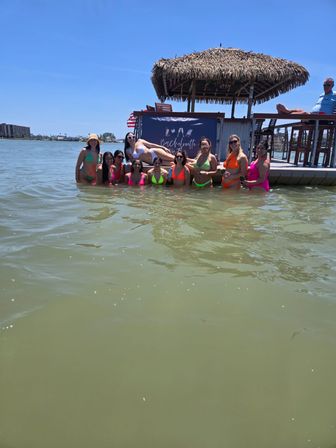 Group of women in bright neon bikinis posing waist-deep in green bay water beside a thatched-roof tiki bar and dock on a sunny bachelorette beach day