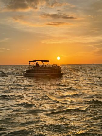 Silhouette of a pontoon boat with people on the ocean at golden-hour sunset, warm orange sky and sun reflecting on rippling water.