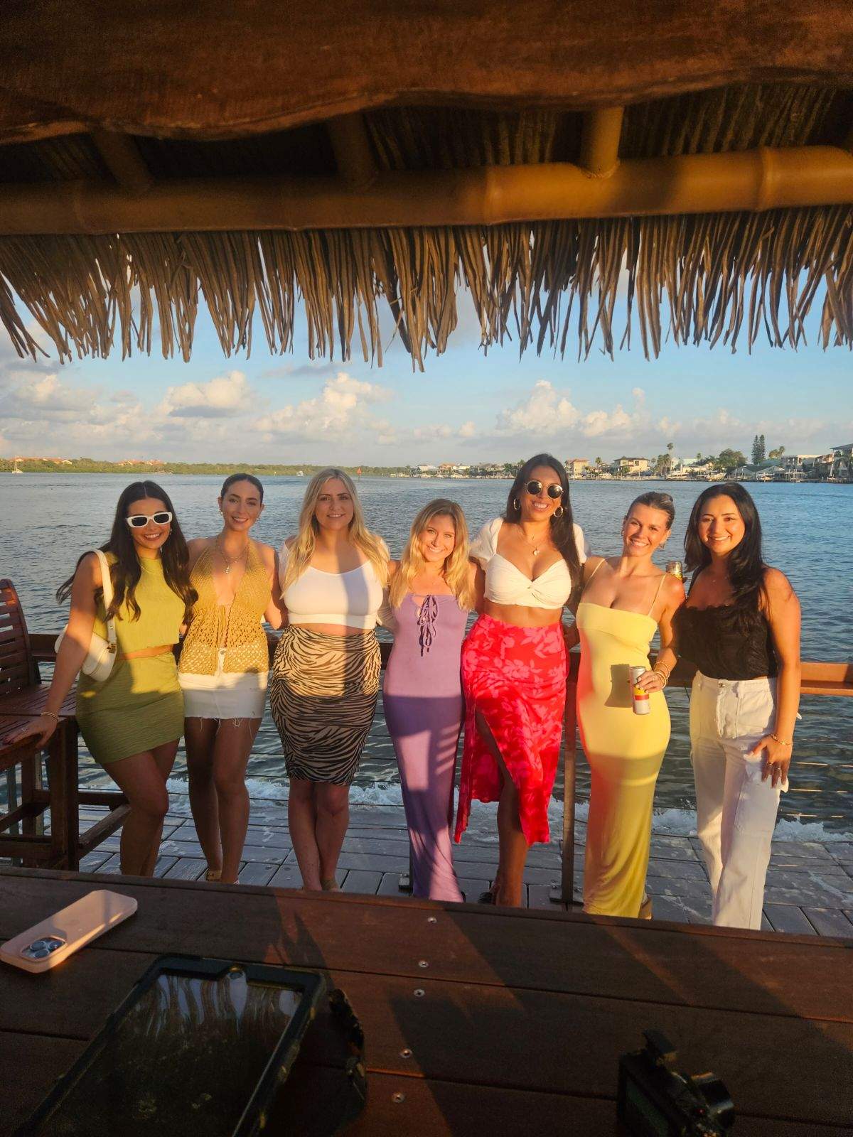 Seven friends in colorful summer outfits posing and smiling on a waterfront thatched-roof dock at sunset, with a calm bay and coastal homes in the background