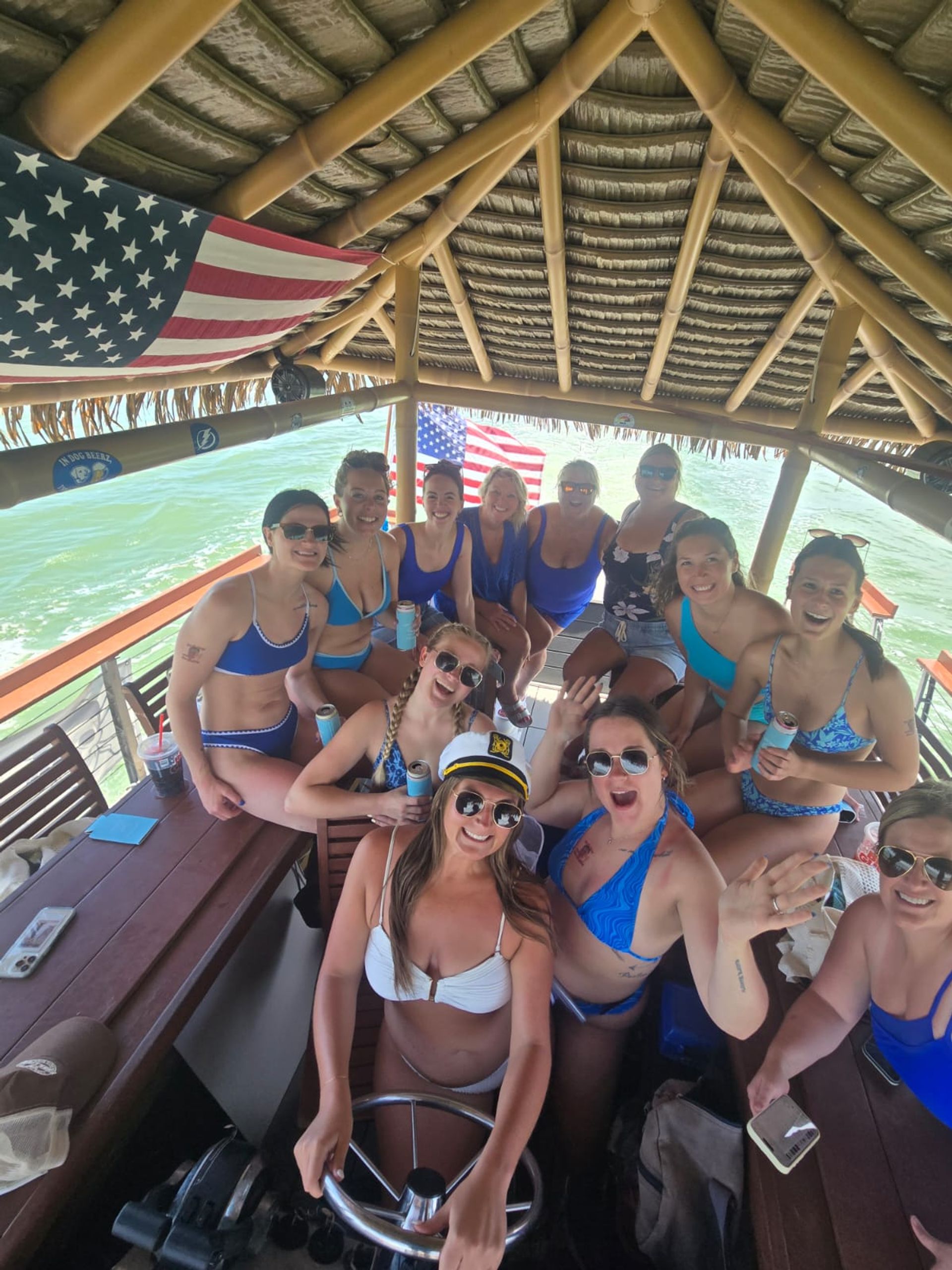 Group of women in swimsuits enjoying a sunny day on a tiki-style boat with a thatched roof and American flags, smiling and waving as one woman steers at the helm wearing a captain’s hat over green coastal water.