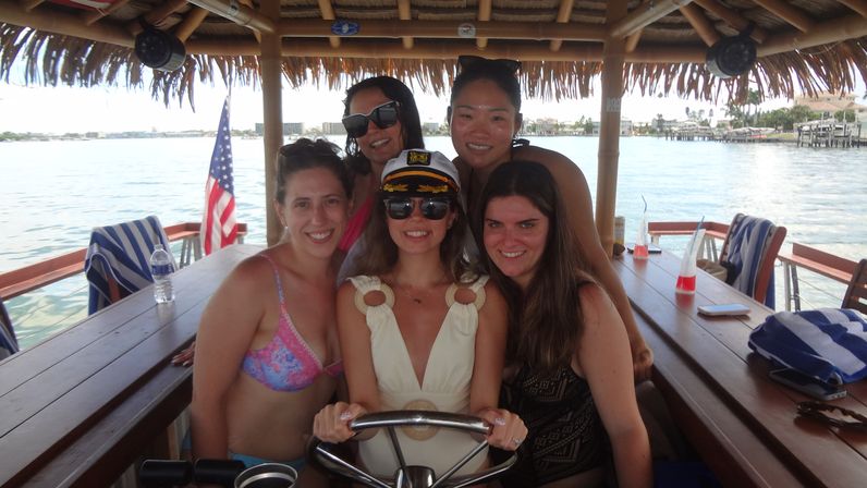 Five friends smiling on a thatched-roof tiki boat over a sunny harbor, a woman in a captain's hat and sunglasses at the helm, American flag, striped beach towels and tropical drinks on the wooden table