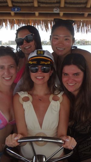 Five friends smiling on a thatched-roof tiki boat over a sunny harbor, a woman in a captain's hat and sunglasses at the helm, American flag, striped beach towels and tropical drinks on the wooden table