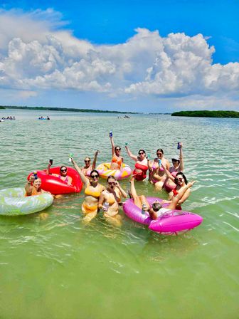 Group of friends in colorful bikinis floating on bright inflatable tubes in shallow green bay water, raising cans and cheering under a blue sky with puffy white clouds and distant mangrove shoreline — sunny beach day.