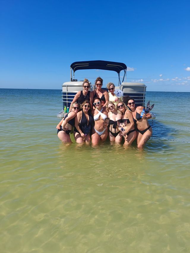 Group of friends in swimsuits posing waist-deep in shallow coastal water in front of an anchored pontoon boat under a clear blue sky, holding a small dog — sunny beach day.