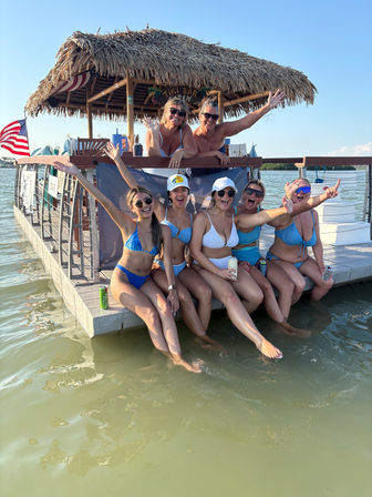 Group of friends in bikinis laughing on a floating tiki-bar dock, legs dangling in shallow bay water under a clear summer sky with an American flag nearby.