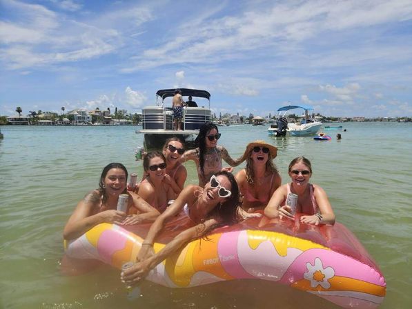 Seven women relaxing on a colorful inflatable float in shallow bay water, surrounded by pontoon boats and waterfront homes on a sunny summer day.