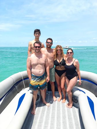Five people smiling together on a pontoon boat in turquoise ocean under a sunny blue sky, wearing swimsuits with a white powerboat passing in the background — bright tropical boat day.