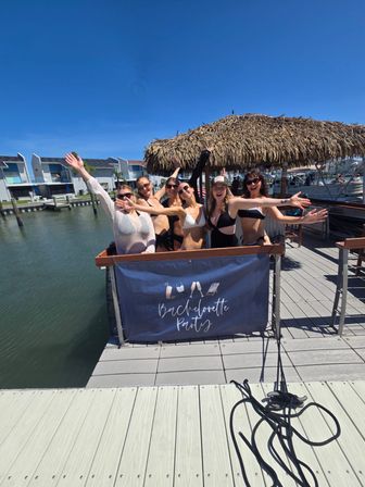 Six women in swimsuits waving on a marina dock under a thatched tiki umbrella with a “Bachelorette Party” banner, boats and waterfront condos in the sunny blue-sky background.