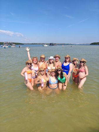 Cheerful group of women in colorful bikinis and straw cowboy hats posing waist‑deep on a sunny shallow sandbar with boats and blue sky in the background