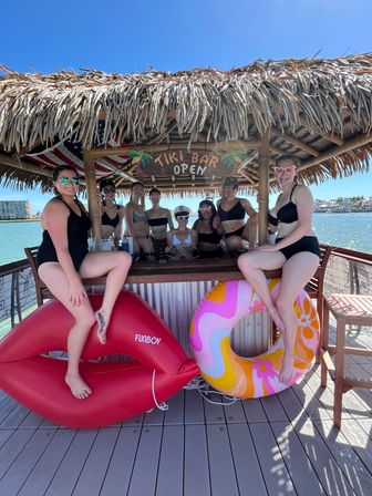 Group of friends in swimsuits at a thatched-roof tiki bar on a sunny waterfront dock with colorful inflatable rings and an "Tiki Bar Open" sign