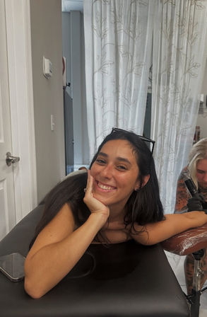 Smiling woman reclining on a tattoo table while a tattoo artist inks her upper arm in a bright studio, sunglasses on her head and phone nearby.