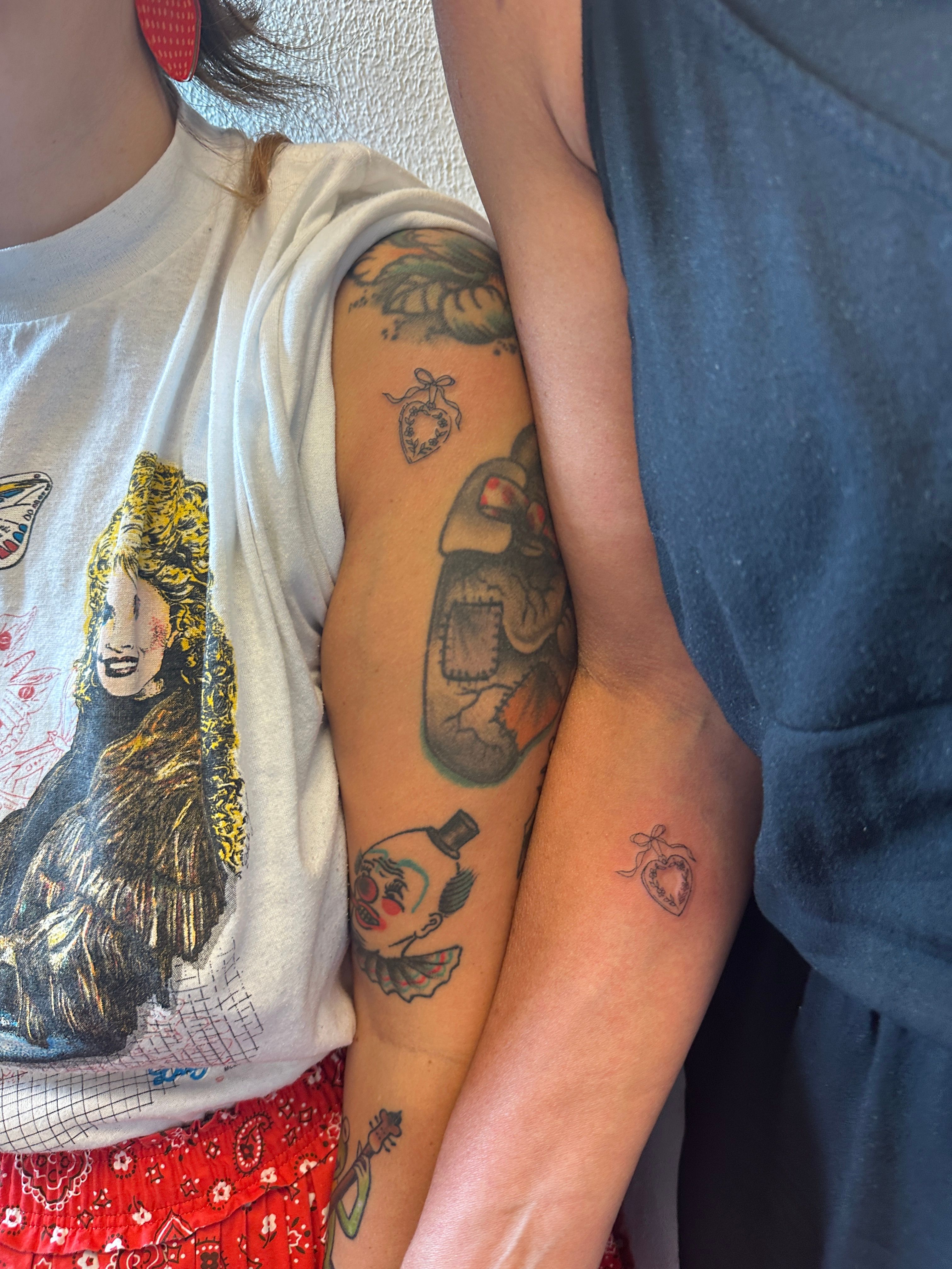 Close-up indoor shot of two forearms side-by-side showing matching tiny heart locket tattoos with ribbon; colorful vintage tattoos (clown, patched heart) visible, one person in a white graphic tee and red bandana skirt, the other in a black top.