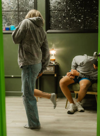 Two people in casual hoodies inside a green-walled tattoo studio — one stands in jeans lifting a sock to show a calf tattoo while the other sits on a bench under a frosted window and vintage lamp.