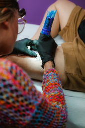 Close-up of a tattoo artist wearing black gloves using a blue-wrapped tattoo machine to ink a client's thigh in a colorful studio setting.