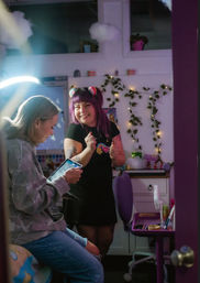 Two friends in a cozy purple-decorated room: one seated using a tablet, the other with purple hair and rainbow clips smiling by a desk with string lights and potted plants.