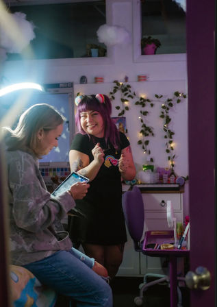 Two friends in a cozy purple-decorated room: one seated using a tablet, the other with purple hair and rainbow clips smiling by a desk with string lights and potted plants.