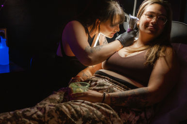 Tattoo artist in gloves using a wrapped tattoo machine on a relaxed, smiling client’s upper chest/collarbone as she reclines holding a drink in a dimly lit tattoo studio