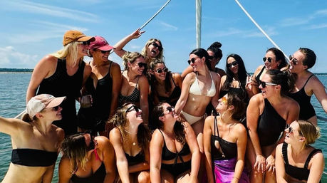 Smiling group of women in swimsuits posing on a sailboat deck under a bright blue sky over calm ocean water — summer boat outing.