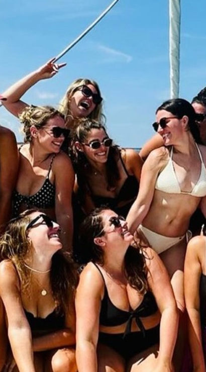 Smiling group of women in swimsuits posing on a sailboat deck under a bright blue sky over calm ocean water — summer boat outing.