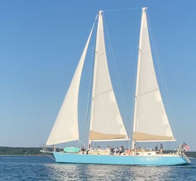 Aqua-hulled two-masted sailing yacht with billowing white sails and passengers cruising calm coastal waters under a clear blue sky, American flag at the stern.