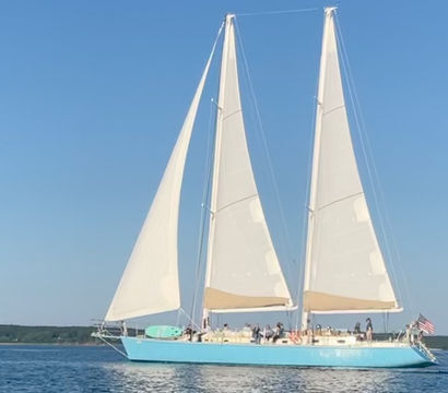 Aqua-hulled two-masted sailing yacht with billowing white sails and passengers cruising calm coastal waters under a clear blue sky, American flag at the stern.
