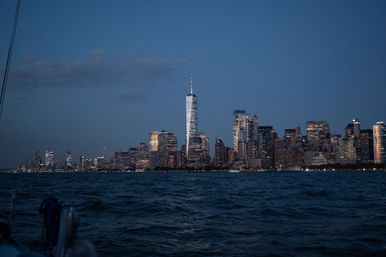 Manhattan skyline at dusk with illuminated One World Trade Center rising above lit skyscrapers, viewed across the Hudson River from a boat