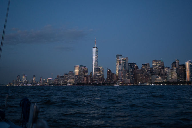 Manhattan skyline at dusk with illuminated One World Trade Center rising above lit skyscrapers, viewed across the Hudson River from a boat